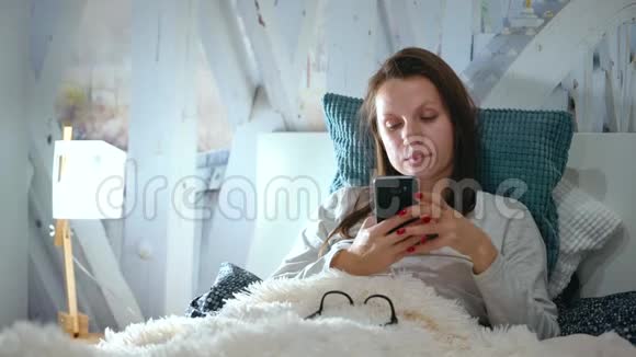 戴眼镜的女人躺在床上睡前使用智能手机视频的预览图
