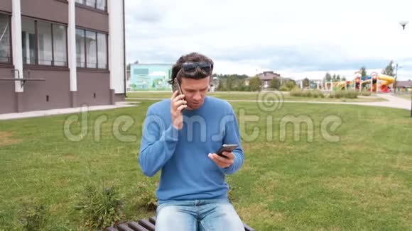 男人拿着两部手机坐在家附近的长凳上两个电话的通话视频的预览图