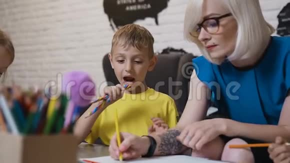 一个穿着黄色t恤带着彩色两支铅笔的小男孩坐在桌子旁看着他的老师画画绘图视频的预览图