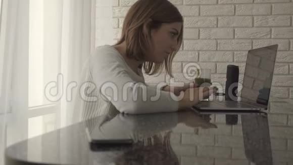 一个在家里用笔记本电脑工作的女人坐在桌子旁喝着健康的绿色冰沙然后伸手去检查她视频的预览图