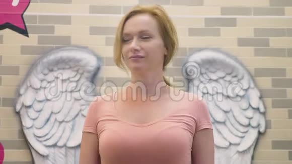 一个金发女人站在墙的背景上以天使的翅膀的形式展开视频的预览图