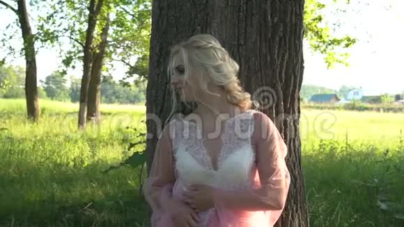 夏天日落时一个穿着粉红色衣服的漂亮女孩靠在一棵大树上化妆的肖像视频的预览图