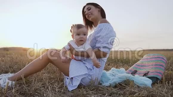 一个穿着浅色连衣裙的年轻女子坐在草地上和一个可爱的婴儿玩耍迷人的肖像视频的预览图