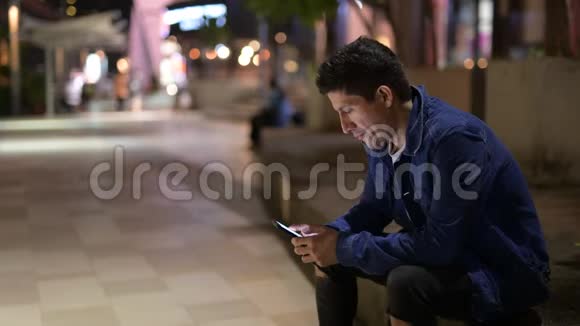 西班牙裔男子晚上坐在街上思考和使用电话视频的预览图