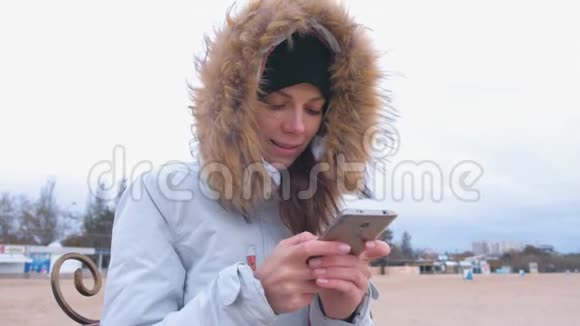 穿着白色羽绒服的女人坐在海滩上的长凳上在手机上输入一条信息视频的预览图
