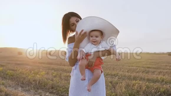 一个戴着大帽子的婴儿在夕阳下和她的母亲在田野里散步的肖像快乐的年轻女人和小儿子在一起视频的预览图