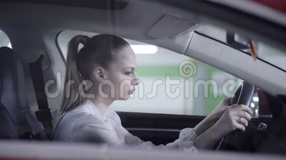 年轻女子穿着白衬衫坐在车里拿着方向盘在车库旁边视频的预览图