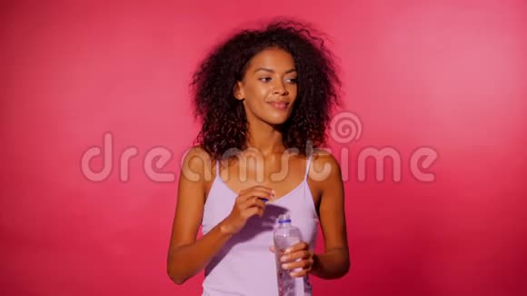 迷人的非洲裔美国运动青年妇女饮用瓶水粉红色墙壁背景穿运动服装的女孩视频的预览图