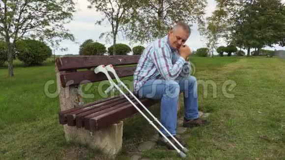坐在长凳上的公园里拄着拐杖的悲伤残疾人视频的预览图