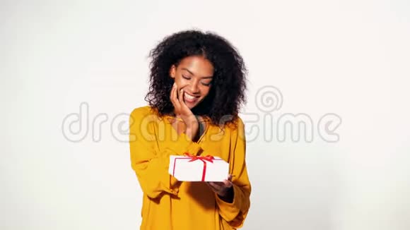 可爱的非裔美国年轻女子穿着黄色的秋装上衣手里拿着带有红色丝带的礼品盒在白色的墙壁背景上鞠躬视频的预览图