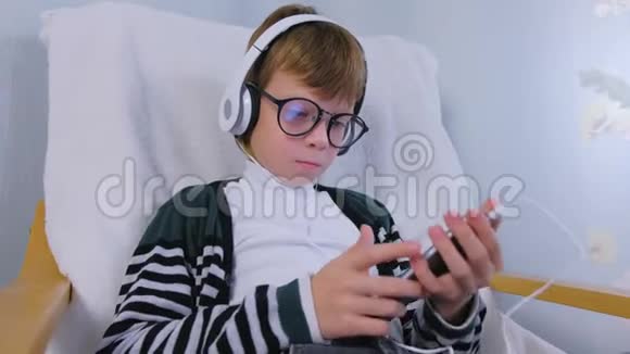 戴眼镜的男孩正坐在家里的扶手椅上听着耳机里的音乐视频的预览图