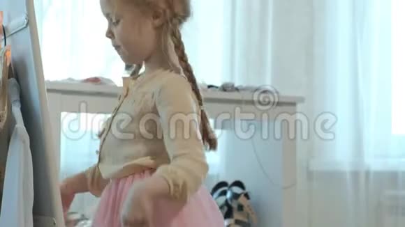 穿着粉红色裙子扎着辫子的开朗的小女孩试着穿着妈妈高跟鞋在镜子前跳舞视频的预览图