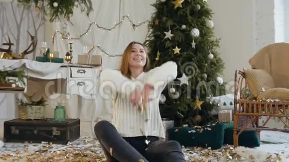 快乐的年轻女子坐在地板上在圣诞树和圣诞树的背景上放着闪闪发光的金色纸屑视频的预览图