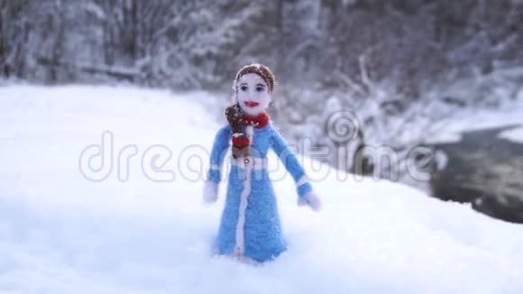 玩具女孩用羊毛做的站在冬天的河流背景下的雪堆里视频的预览图