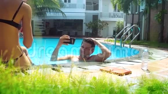 男人在泳池里自拍穿比基尼的苗条女孩坐在障碍上视频的预览图