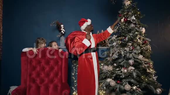 当穿着圣诞老人服装的非洲男人把玩具放在圣诞老人身上时小可爱的女孩偷偷地躲在椅子后面视频的预览图