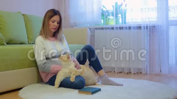 穿着精致的白色粉红色夹克和牛仔裤的漂亮年轻女人坐在房间里的米色圆地毯上抚摸着巨大的白色红猫视频的预览图