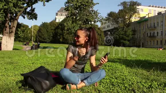 美丽的年轻女孩戴着太阳镜坐在公园的草地上打开手机里她最喜欢的旋律视频的预览图