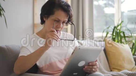 成熟的家庭主妇有时间与平板电脑坐在沙发在家里的内部视频的预览图