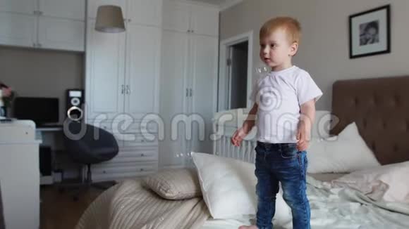 一个穿着白色T恤和蓝色牛仔裤的男孩在父母卧室的床上看到肥皂泡视频的预览图