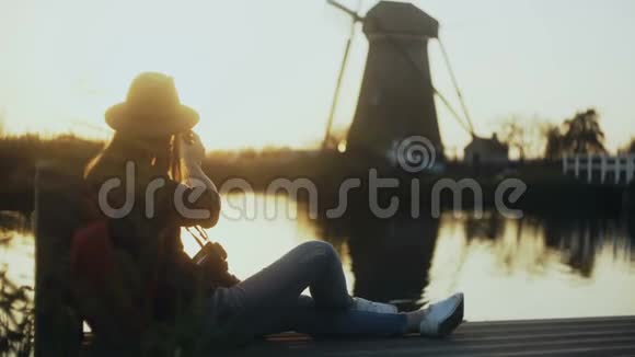 戴帽子的女人坐在日落湖码头荷兰拥有智能手机和相机的女性博主拍了一张风车照片4K视频的预览图