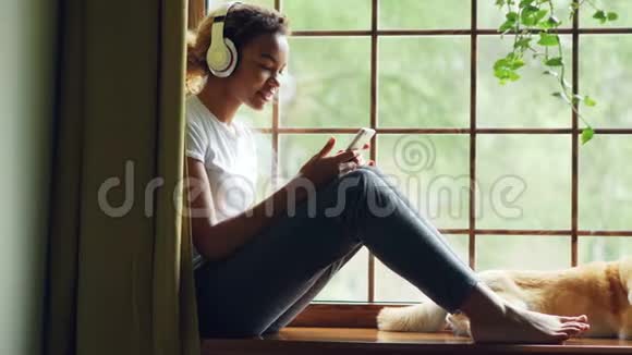 漂亮的非裔美国女孩正在用耳机听音乐用智能手机坐在窗台上视频的预览图