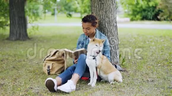漂亮的非裔美国女学生正坐在草坪上的公园里看书而她那只有教养的狗正坐在她身边视频的预览图