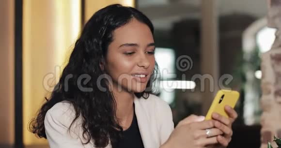 快乐的年轻女商人用黄色智能手机在社交媒体上聊天坐在咖啡馆里微笑的年轻女性用户视频的预览图