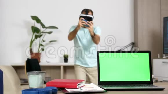 男人玩虚拟现实电子游戏而桌子旁的女人一边看着绿色屏幕电脑一边在上面聊天视频的预览图