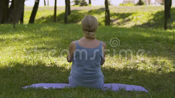 女性坐在垫子上做瑜伽体式在公园冥想莲花姿势慢动作视频的预览图