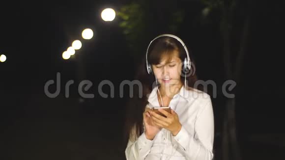 戴着耳机的女人听着歌舞通过触摸电话在城市夜间公园唱歌慢动作视频的预览图