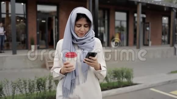 戴着灰色头巾的年轻女子喝着咖啡看着智能手机视频的预览图