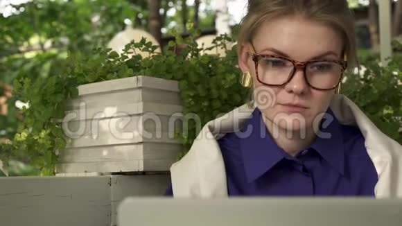 戴眼镜的嬉皮士女孩在她笔记本电脑附近的夏日咖啡馆喝咖啡视频的预览图