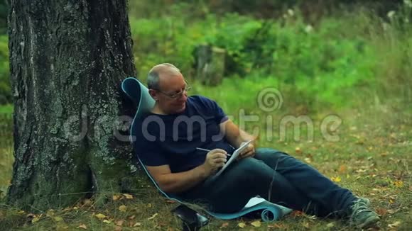 一个戴眼镜的人坐在公园的树下用铅笔在笔记本上写字视频的预览图