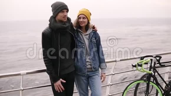 一对年轻的幸福夫妇站在一起在海边拥抱穿暖和的衣服戴冷帽的年轻人视频的预览图