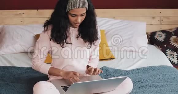 快乐的漂亮女孩穿着粉红色睡衣坐在房子里的床上在笔记本电脑上工作她在微笑视频的预览图