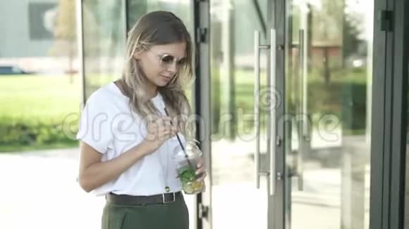 戴墨镜的金发美女喝着冰冷的莫希托在公园散步视频的预览图