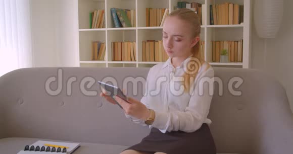 年轻漂亮的白种人金发女学生用平板电脑坐在室内沙发上的特写照片视频的预览图