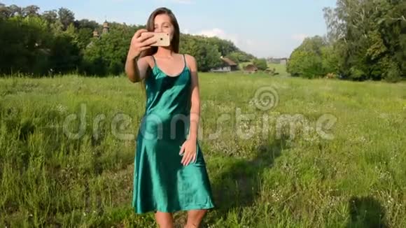 美丽快乐的年轻女性穿着夏日连衣裙在农村背景下用智能手机自拍视频的预览图