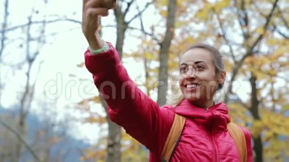穿着红色夹克和小背包的漂亮女徒步旅行者正站在黄色的秋林背景上用电话视频的预览图