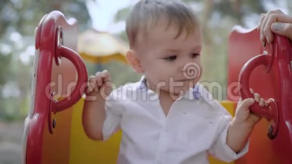 一个可爱的小男孩穿着新鲜的白色衬衫在操场上摇摆的肖像视频的预览图