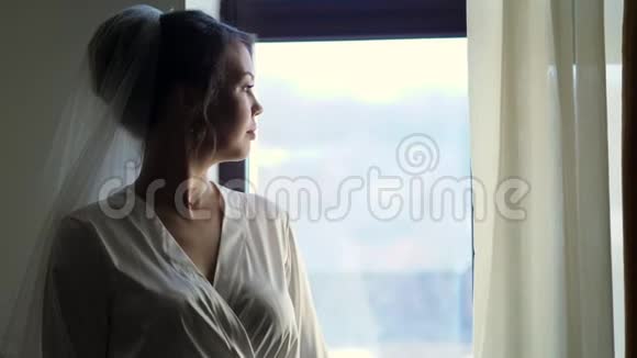 一个新娘的肖像一个美丽的女孩戴着面纱穿着白色长袍望着窗外视频的预览图