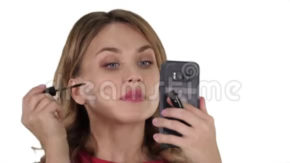 女人在睫毛上涂黑色睫毛膏在白色背景上看着她的手机视频的预览图