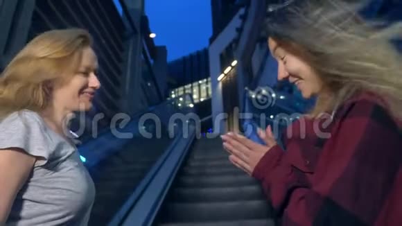 两个女孩亚洲人和高加索人晚上在现代城市的街道上聊天站在自动扶梯上视频的预览图