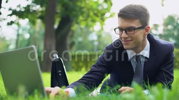 成功的年轻经理躺在草地上微笑着在笔记本电脑上聊天视频的预览图