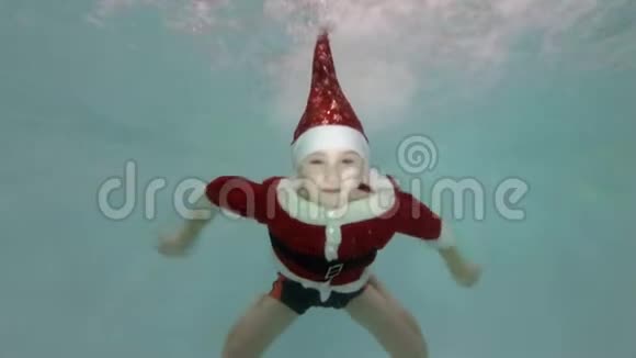 快乐的小男孩穿着红色西装圣诞老人在水中的急流中在游泳池里游泳微笑着看着镜头视频的预览图