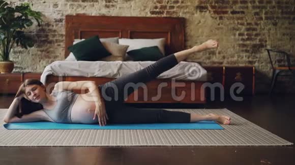漂亮的怀孕女孩正在做体育锻炼抬起腿躺在卧室的地板上瑜伽垫上有阁楼风格的房间和视频的预览图