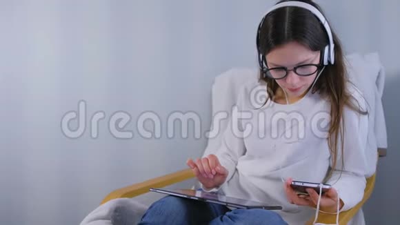 戴眼镜和耳机的女人在网上商店里用智能手机和平板电脑坐在扶手椅上购物视频的预览图