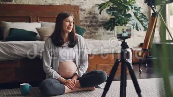 快乐的怀孕女孩正在用三脚架上的智能手机为她的在线视频制作视频女孩正在和订户交谈视频的预览图