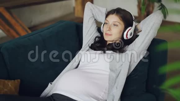 期待妈妈的美丽女孩正在听音乐戴着耳机在家里的沙发上放松享受休息和休息视频的预览图